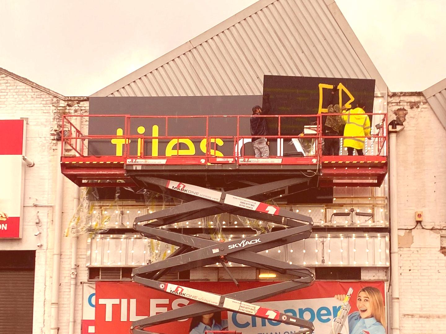 Installation team working on Tiles DIY storefront signage using scaffolding and lifts.